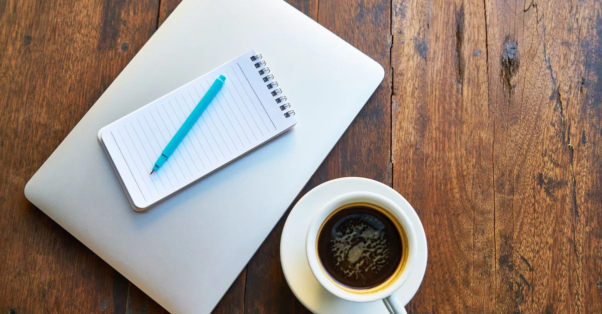 Vue de dessus d'une tasse à café, d'un bloc-notes, d'un stylo bleu et d'un ordinateur portable sur un bureau en bois.