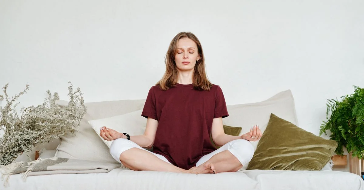 Femme méditant à l'intérieur sur un sofa, dégageant calme et pleine conscience, entourée de verdure.